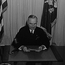 President Harry S. Truman seated at a desk, before a microphone, announcing the end of World War II in Europe