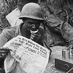 Private First Class Clarence Whitmore, voice radio operator, 24th Infantry Regiment, reads the latest news while enjoying chow during lull in battle, near Sangju, Korea.